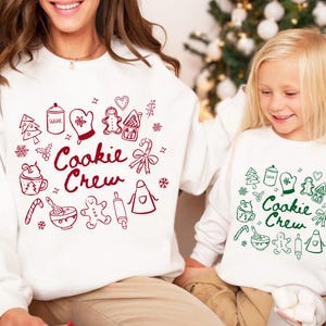 May include: Two white sweatshirts with long sleeves, featuring a red and green "Cookie Crew" design. The design includes illustrations of baking-related items like a gingerbread man, rolling pin, and sugar jar. The sweatshirts are worn by a person and a child.