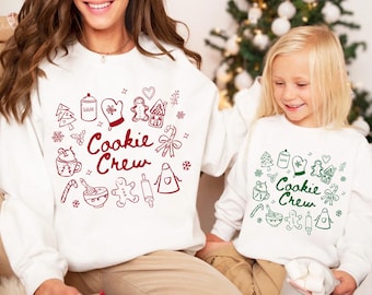 May include: Two white sweatshirts with long sleeves, featuring a red and green "Cookie Crew" design. The design includes illustrations of baking-related items like a gingerbread man, rolling pin, and sugar jar. The sweatshirts are worn by a person and a child.