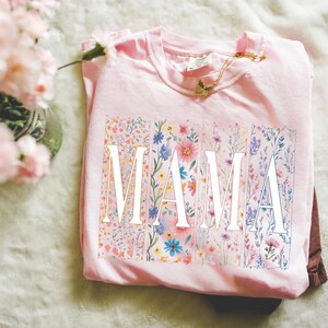 May include: A light pink t-shirt with the word "MAMA" in white block letters. Each letter is filled with a different floral pattern in shades of pink, blue, and yellow. A gold butterfly necklace rests on the shirt.
