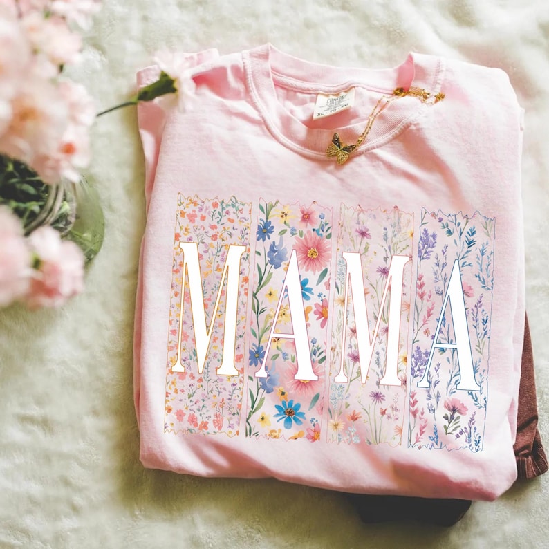 May include: A light pink t-shirt with the word "MAMA" in white block letters. Each letter is filled with a different floral pattern in shades of pink, blue, and yellow. A gold butterfly necklace rests on the shirt.