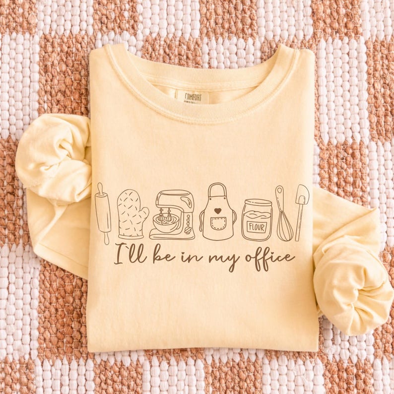 May include: A pale yellow long-sleeved top featuring the phrase "I'll be in my office" and a line drawing of baking equipment. The illustration includes a rolling pin, oven glove, mixer, apron, flour jar, whisk, and spatula.