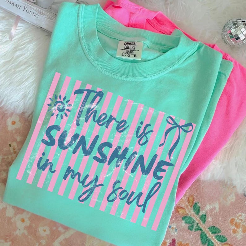 Puede incluir: Una camiseta Comfort Colors verde agua con el texto "There is SUNSHINE in my soul" en escritura azul sobre un dise&ntilde;o de rayas rosas. Una camiseta rosa intenso es visible en el fondo. La camiseta tiene cuello redondo y mangas cortas.