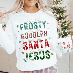 May include: White sweatshirt with the text "dance like FROSTY, shine like RUDOLPH, give like SANTA, love like JESUS" in green, brown, and red. The sweatshirt has snowflakes around the text and is perfect for the holiday season.