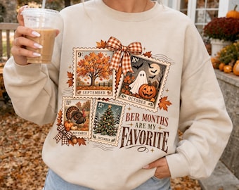 May include: A beige crewneck sweatshirt featuring a graphic print of four postage stamp-style illustrations representing September, October, November, and December, topped with a plaid bow and the text "BER MONTHS ARE MY FAVORITE."