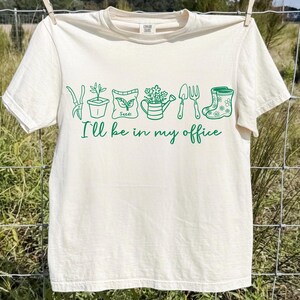Puede incluir: Camiseta color crema con dibujos verdes de herramientas de jardiner&iacute;a y plantas. El texto "I'll be in my office" est&aacute; escrito en verde debajo. La camiseta est&aacute; colgada en un tendedero.