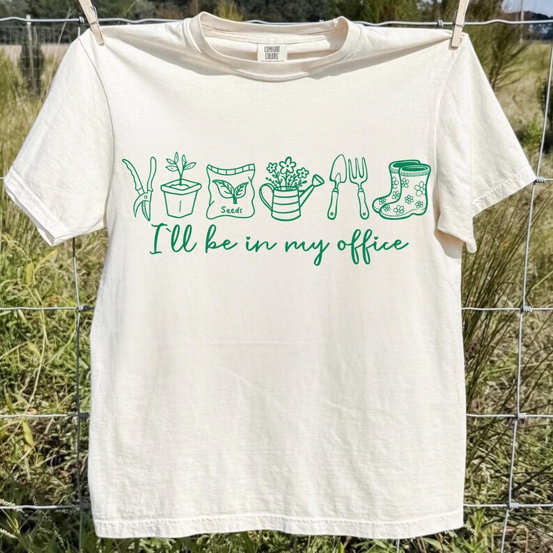 Puede incluir: Camiseta color crema con dibujos verdes de herramientas de jardiner&iacute;a y plantas. El texto "I'll be in my office" est&aacute; escrito en verde debajo. La camiseta est&aacute; colgada en un tendedero.