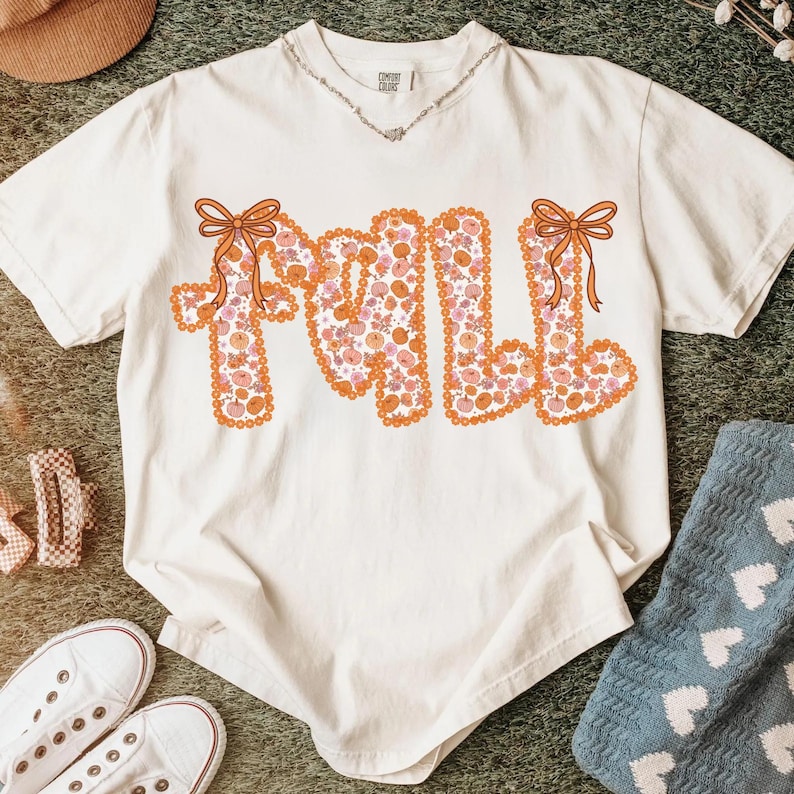 May include: Cream-colored t-shirt with the word "FALL" in orange, floral-patterned letters. Each letter is outlined in orange and topped with an orange bow. The shirt is part of a fall-themed collection.