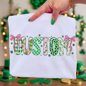 May include: White t-shirt with the word "CUSTOM" in a festive design. The letters feature green and pink patterns, including shamrocks, checkered patterns, and leopard print. Pink bows accent the beginning and end of the word.