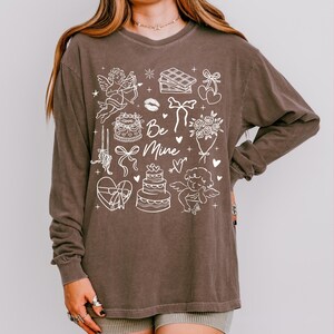 May include: A brown long-sleeve shirt with a white graphic design featuring cherubs, cakes, hearts, and the text "Be Mine." The shirt has a relaxed fit and is made of a soft material, perfect for Valentine's Day or everyday wear.