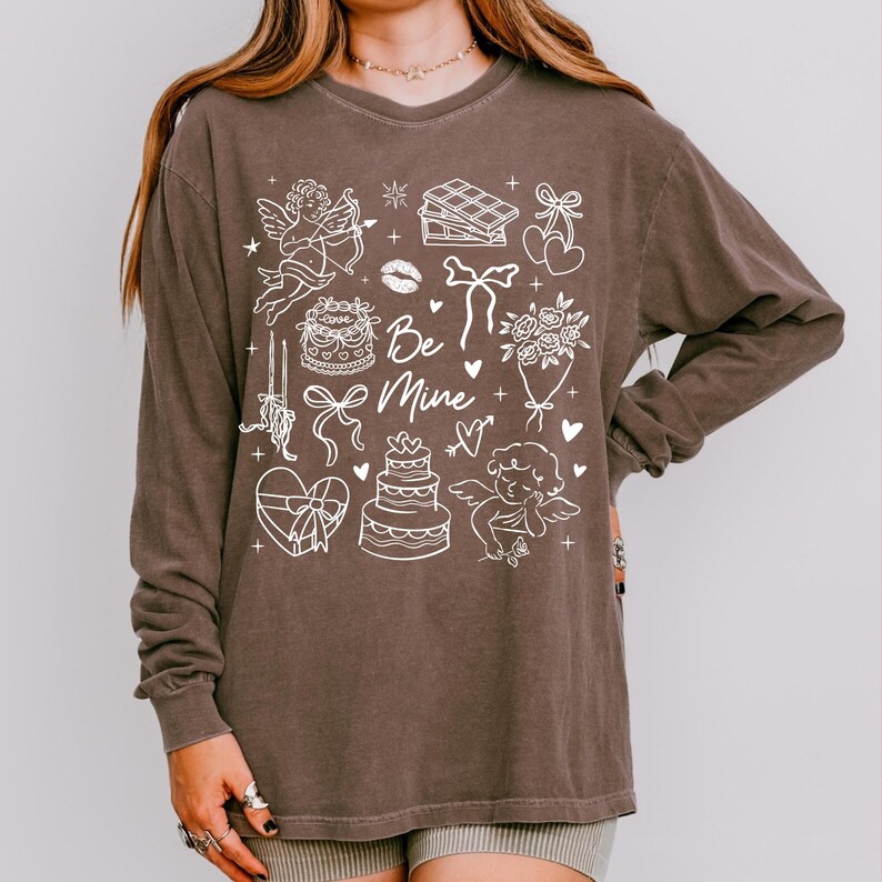 May include: A brown long-sleeve shirt with a white graphic design featuring cherubs, cakes, hearts, and the text "Be Mine." The shirt has a relaxed fit and is made of a soft material, perfect for Valentine's Day or everyday wear.