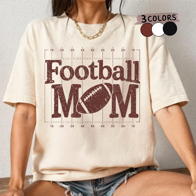 May include: Cream-colored t-shirt with the words "Football MOM" in a distressed brown font, along with a football graphic. The design includes yard lines and numbers. Three color options are shown in the upper right corner.