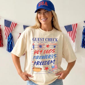 以下が含まれることがあります： ホットドッグ、花火、そして「Hot Dogs Fireworks Freedom」という言葉が描かれた、ゲストチェックのグラフィックが特徴のクリーム色のTシャツ。シャツには青と赤のアメリカ国旗のデザインも施されています。「USA」と書かれた青い野球帽が着用されています。