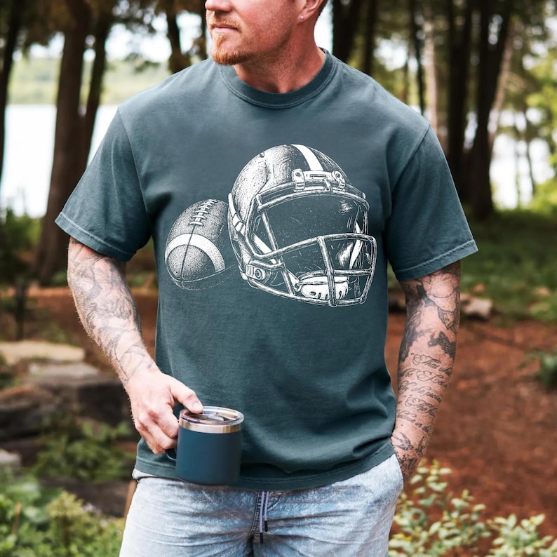 May include: A dark teal short-sleeve t-shirt featuring a white graphic illustration of an American football helmet and a football. The design is centered on the chest of the shirt, showcasing detailed line work of the sports equipment.