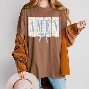 May include: Brown t-shirt with the word "AMEN" in light blue letters. The letters are set against a floral background. A light blue bow is beneath the word. The shirt is worn with a brown jacket and blue jeans.
