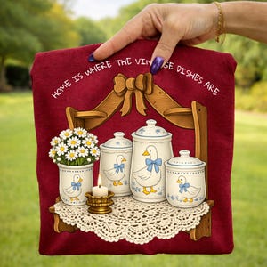 Op de afbeelding: Een bordeauxrode theedoek met de tekst "HOME IS WHERE THE VINTAGE DISHES ARE". De theedoek toont een plank met een vaas met madeliefjes, een kaars en keramische potten met eendenontwerpen.