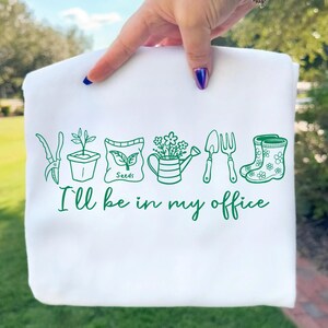 Puede incluir: Sudadera blanca con un dise&ntilde;o verde con tem&aacute;tica de jard&iacute;n. El dise&ntilde;o incluye herramientas de jardiner&iacute;a, una regadera y el texto "I'll be in my office."
