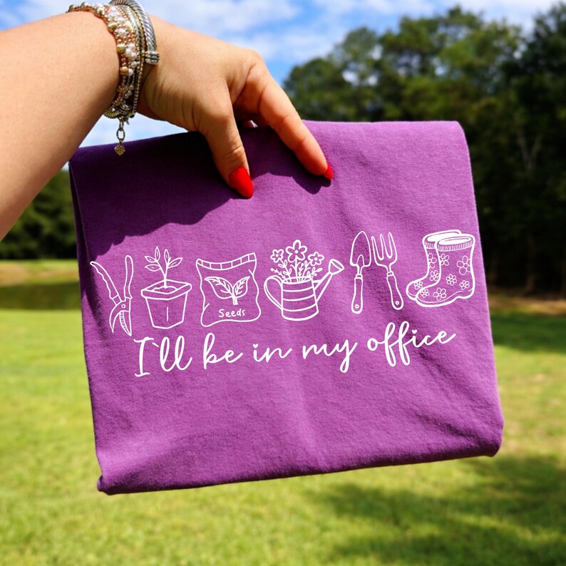 Puede incluir: Una camiseta morada con ilustraciones blancas de jardiner&iacute;a y el texto "I'll be in my office". El dise&ntilde;o incluye una paleta, una regadera, botas y un paquete de semillas.