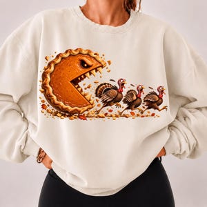 May include: A cream-colored crewneck sweatshirt featuring a graphic print of a large, orange pumpkin pie with sharp teeth chasing three brown turkeys. The pie has a crusty edge and is surrounded by scattered autumn leaves and pie crust crumbs.