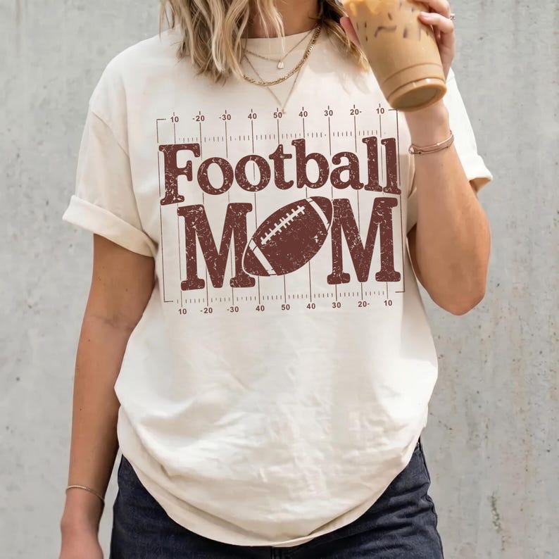 May include: Cream-colored t-shirt with the words "Football MOM" in a distressed brown font, along with a football graphic. The design includes a grid pattern with yard line markings. The shirt has rolled-up sleeves.