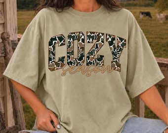 May include: A sage green short-sleeve t-shirt featuring the text COZY in large block letters with a green, brown, and tan camouflage pattern, and the word season written in a tan cursive script font centered below the block letters.