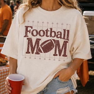 May include: Cream-colored t-shirt with a maroon football graphic. The text reads "Football MOM" with a football image in the center. The design includes yard lines and numbers.
