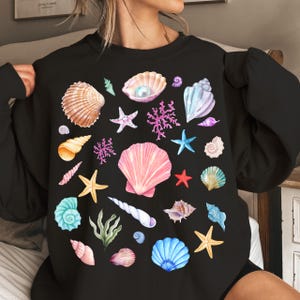 May include: Black sweatshirt with a colorful seashell print. The print features various seashells, starfish, and coral in shades of pink, blue, green, and yellow.