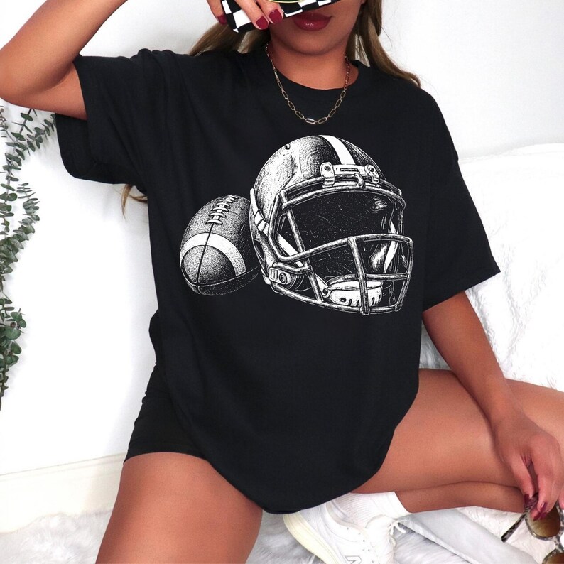 May include: A black short-sleeved t-shirt featuring a white graphic print of an American football helmet and a football. The design is rendered in a detailed, sketch-like style with visible shading and lines, centered on the front of the garment.