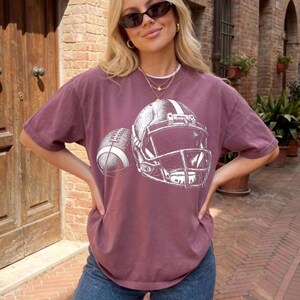 May include: A maroon short-sleeve t-shirt featuring a white graphic print of a football helmet and a football. The shirt has a crew neckline and a relaxed fit. The graphic is centered on the chest, showcasing detailed line work of the sports gear.