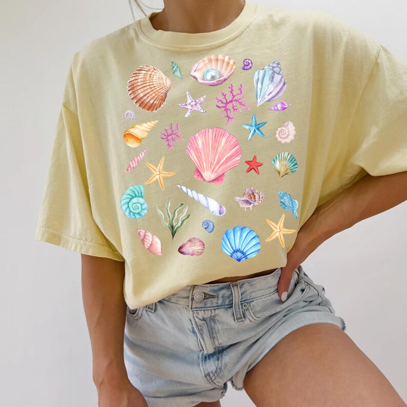 May include: A yellow t-shirt with a colorful seashell and starfish design. The shirt has a relaxed fit and short sleeves.