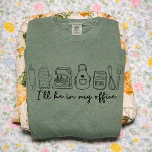 May include: Sage green crew neck sweatshirt featuring a black line drawing of baking tools and the text "I'll be in my office." The design includes a rolling pin, oven mitt, mixer, apron, flour jar, whisk, and spatula. The sweatshirt is folded on floral fabric.