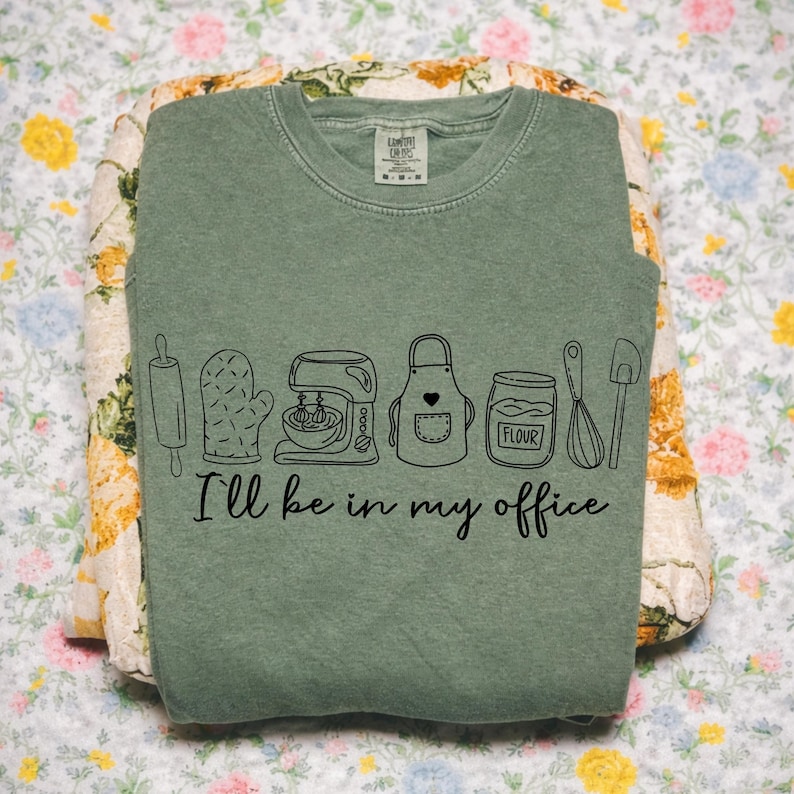 May include: Sage green crew neck sweatshirt featuring a black line drawing of baking tools and the text "I'll be in my office." The design includes a rolling pin, oven mitt, mixer, apron, flour jar, whisk, and spatula. The sweatshirt is folded on floral fabric.