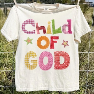 Op de afbeelding: Crèmekleurig T-shirt met de tekst "CHILD OF GOD" in kleurrijke letters in patchwork-stijl. De letters zijn in verschillende patronen, waaronder ruitjes en stippen. Twee sterren zijn ook in het ontwerp opgenomen. Het shirt hangt aan een waslijn.