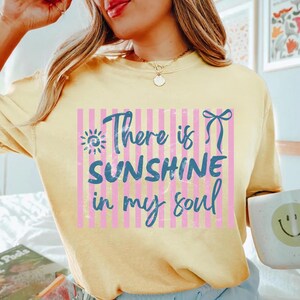 Puede incluir: Sudadera amarilla clara con la frase "There is SUNSHINE in my soul" en azul. El texto est&aacute; sobre un fondo de rayas rosas y blancas. Un sol y un lazo acent&uacute;an el texto.