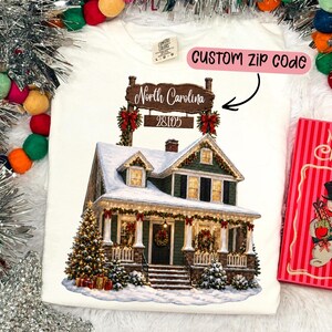 May include: A white t-shirt featuring a graphic of a two-story house covered in snow with Christmas decorations. Above the house is a wooden sign reading North Carolina and 28105. A pink text box points to the sign with the text CUSTOM ZIP CODE.