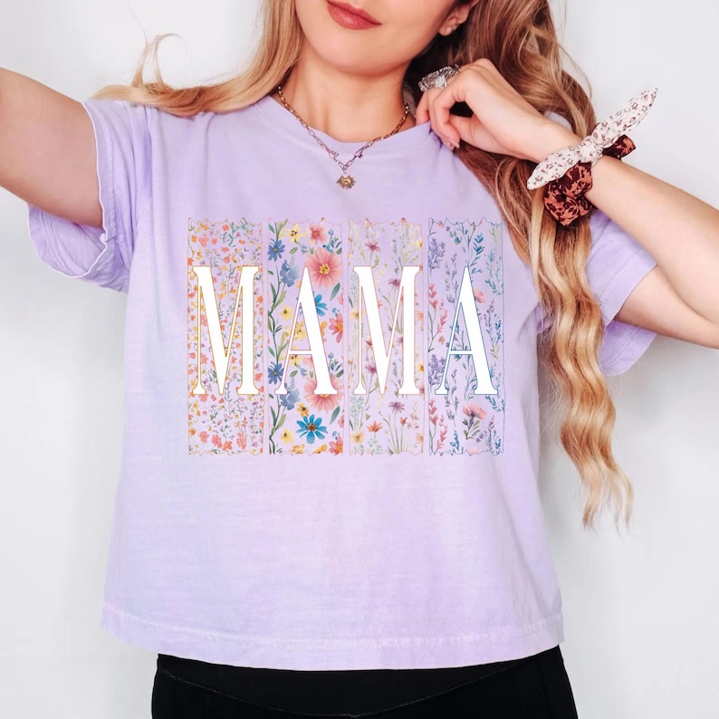 May include: A light purple t-shirt with the word "MAMA" in white block letters. Each letter is overlaid on a vertical floral pattern in shades of pink, blue, and yellow. The shirt has short sleeves and a crew neck.