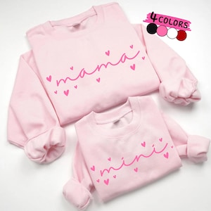 May include: Two pale pink sweatshirts, one adult and one child size, with the words "mama" and "mini" in pink script, decorated with pink hearts. A colour palette of black, pink, white, and red is also shown.