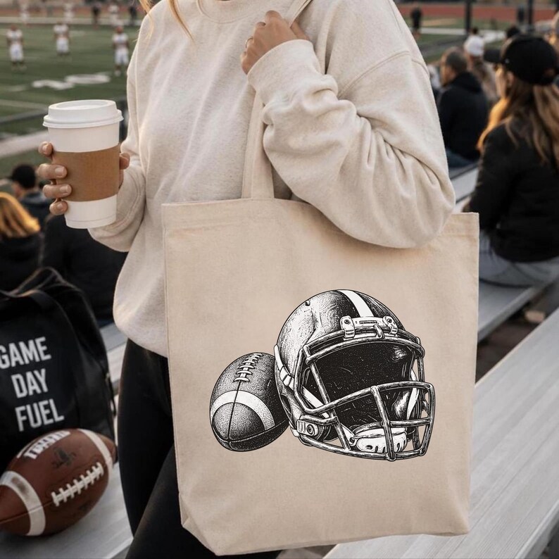 May include: A beige canvas tote bag featuring a black and white ink-style illustration of an American football helmet positioned next to an American football. The design is centered on the front of the bag, showcasing detailed shading and lines.