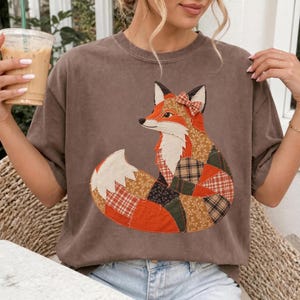May include: A brown short-sleeved t-shirt featuring a large, central graphic of a fox. The fox is constructed from various fabric patches in shades of orange, brown, and green with plaid and floral patterns, and wears a small plaid bow on its head.