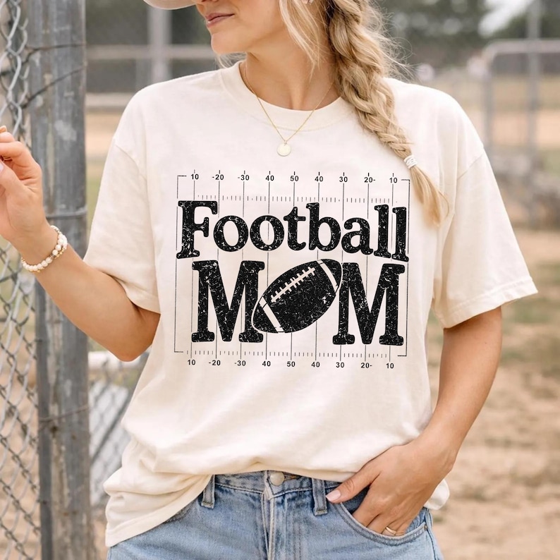 May include: Cream-colored t-shirt with the text "Football MOM" in black, featuring a football graphic. The design includes yard lines. The shirt is a casual style.