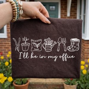 Puede incluir: Un bolso tote marr&oacute;n oscuro con dibujos lineales blancos de herramientas de jardiner&iacute;a y plantas. El texto "I'll be in my office" est&aacute; debajo de las ilustraciones. El bolso lo sostiene una persona.