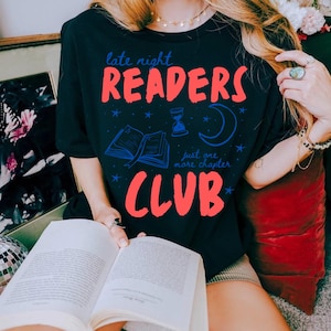 May include: Black t-shirt with the text "late night READERS CLUB" in red, with blue accents of a book, moon, hourglass, and stars. The shirt is being worn while holding a book.