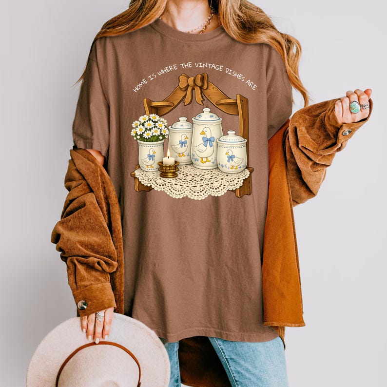 Op de afbeelding: Bruin t-shirt met een afbeelding van een plank met vintage servies, een vaas met bloemen en een brandende kaars. De tekst "HOME IS WHERE THE VINTAGE DISHES ARE" staat boven de plank. Het t-shirt wordt door een persoon gedragen.