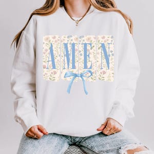 May include: White sweatshirt with the word "AMEN" in light blue block letters. The letters are set against a floral background with a light blue bow at the bottom. The sweatshirt is a casual style.