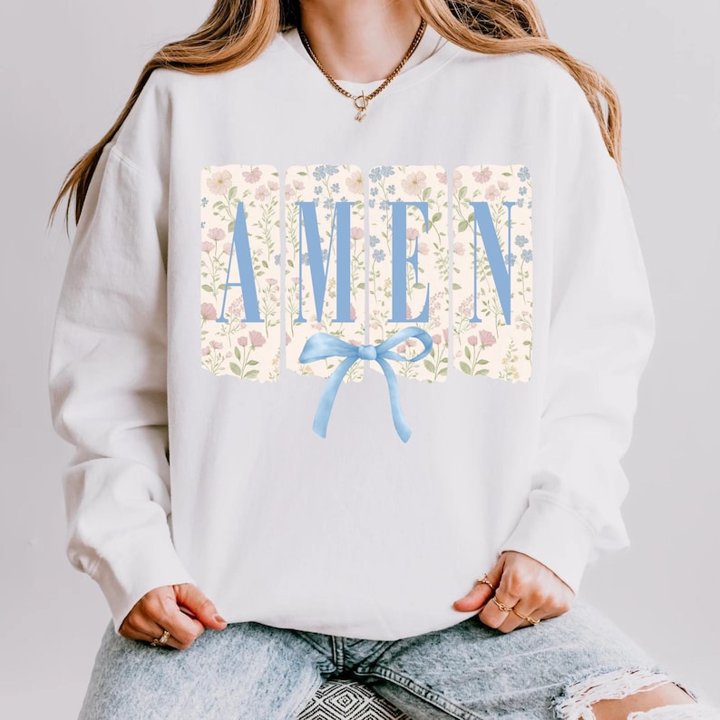 May include: White sweatshirt with the word "AMEN" in light blue block letters. The letters are set against a floral background with a light blue bow at the bottom. The sweatshirt is a casual style.
