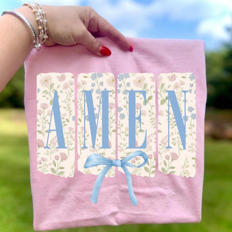 May include: A light pink t-shirt with the word "AMEN" in light blue block letters. The letters are set against a floral background with pink and blue flowers. A light blue bow is below the word.