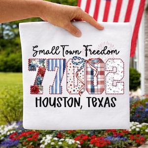 以下が含まれることがあります： 「Small Town Freedom 7702 Houston, Texas」の文字が入った白い正方形のクッション。数字は赤、白、青の花柄、ストライプ柄、星柄で装飾されています。手でクッションを持っています。