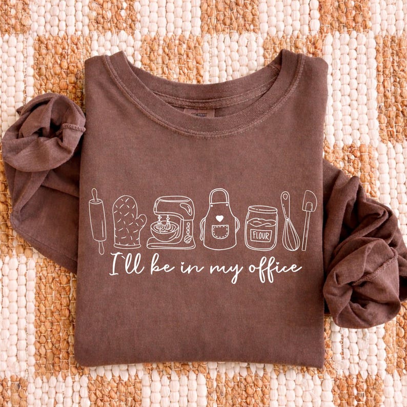 May include: A brown long-sleeved top featuring a white line drawing of baking equipment and the words "I'll be in my office". The design includes a rolling pin, oven glove, mixer, apron, flour jar, whisk, and spatula.