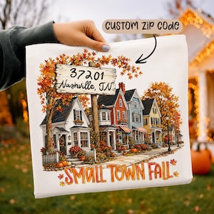 Puede incluir: Una bolsa de tela blanca con una colorida ilustración otoñal de una hilera de casas con árboles naranjas y amarillos. Un letrero de madera muestra el texto 37201 Nashville, TN. El texto inferior dice SMALL TOWN FALL en letras naranjas y rojas.