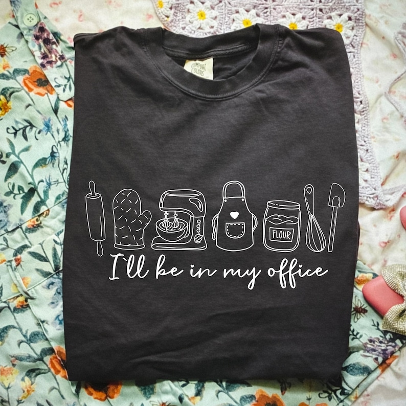 May include: Black t-shirt featuring a white outline of baking equipment: a rolling pin, oven glove, mixer, apron, flour jar, whisk, and spatula. The text "I'll be in my office" is printed below the illustrations.