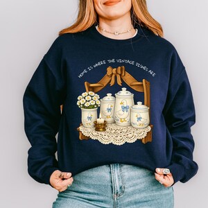 Op de afbeelding: Marineblauwe sweatshirt met een grillige illustratie van een plank met vintage servies, een vaas met bloemen en een kaars. De tekst "HOME IS WHERE THE VINTAGE DISHES ARE" staat boven de plank. Het ontwerp bevat eenden met blauwe strikken.